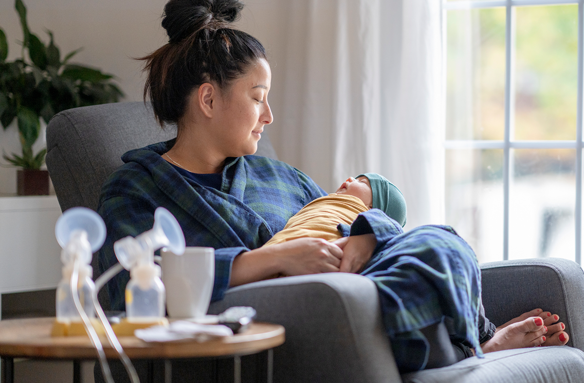 A new mother sitting on a rocking chair in her living room wearing a housecoat holding her newborn baby. There is a Spectra breast pump and nursing supplies around.